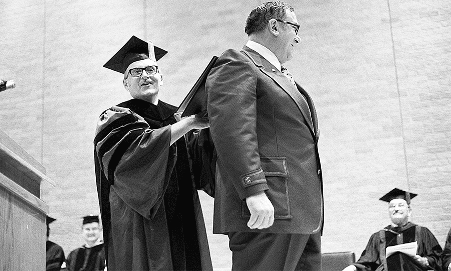 President Flawn reenacts a historic moment—the signing of legislation that created UTSA—by signing the first diploma against Rep. Frank Lombardino’s back.