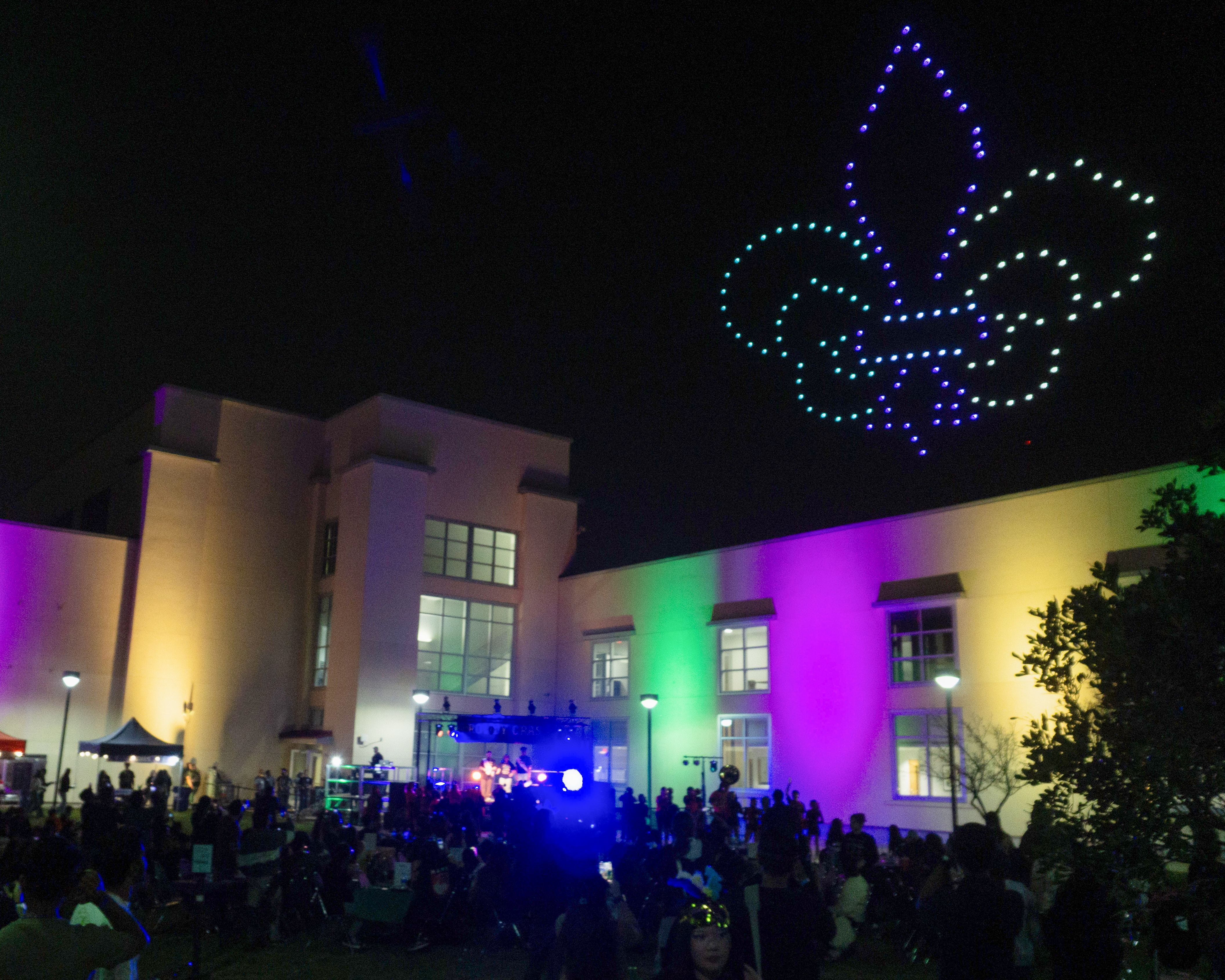 A crowd gathers at night near a building with yellow, purple and green projected on the walls and while drones create a yellow purple and green Fleur-de-lis in the sky.
