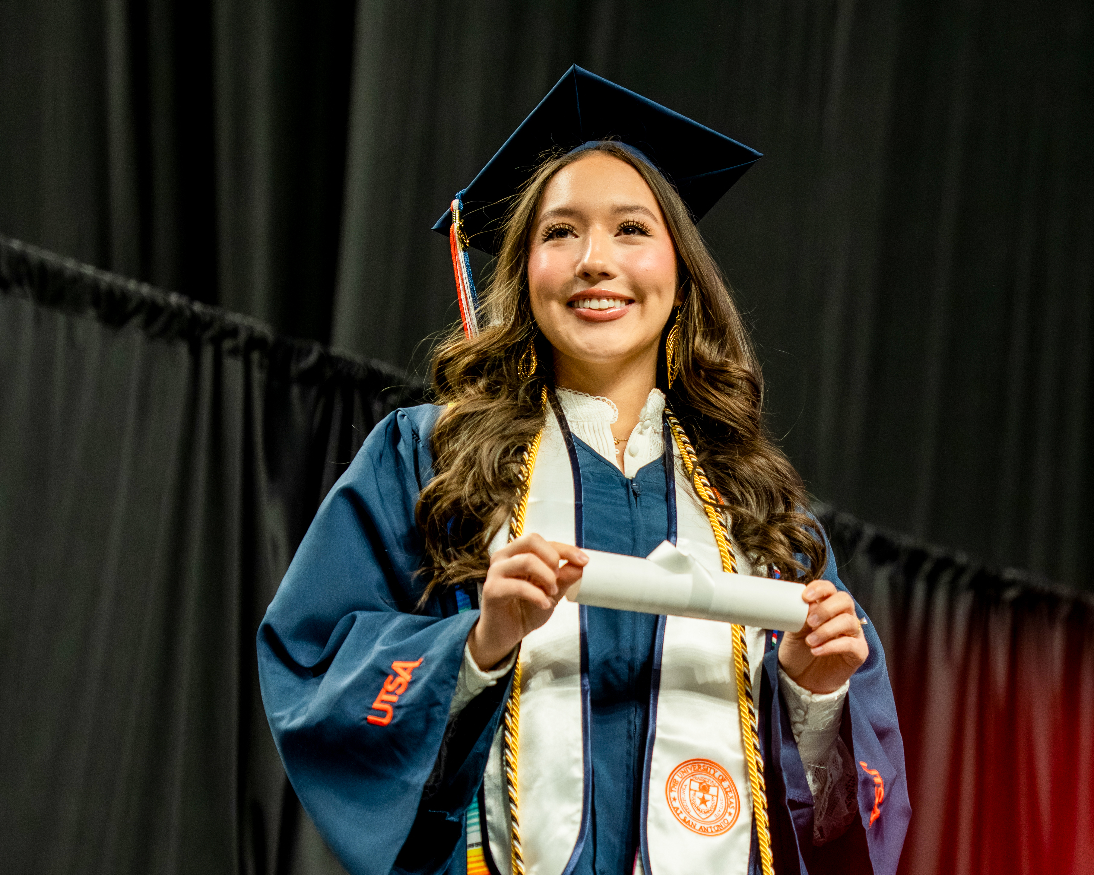A student dresses in a graduation gown and cap with cords and stolls wrapped around her neck holds on to a graduation scroll. 