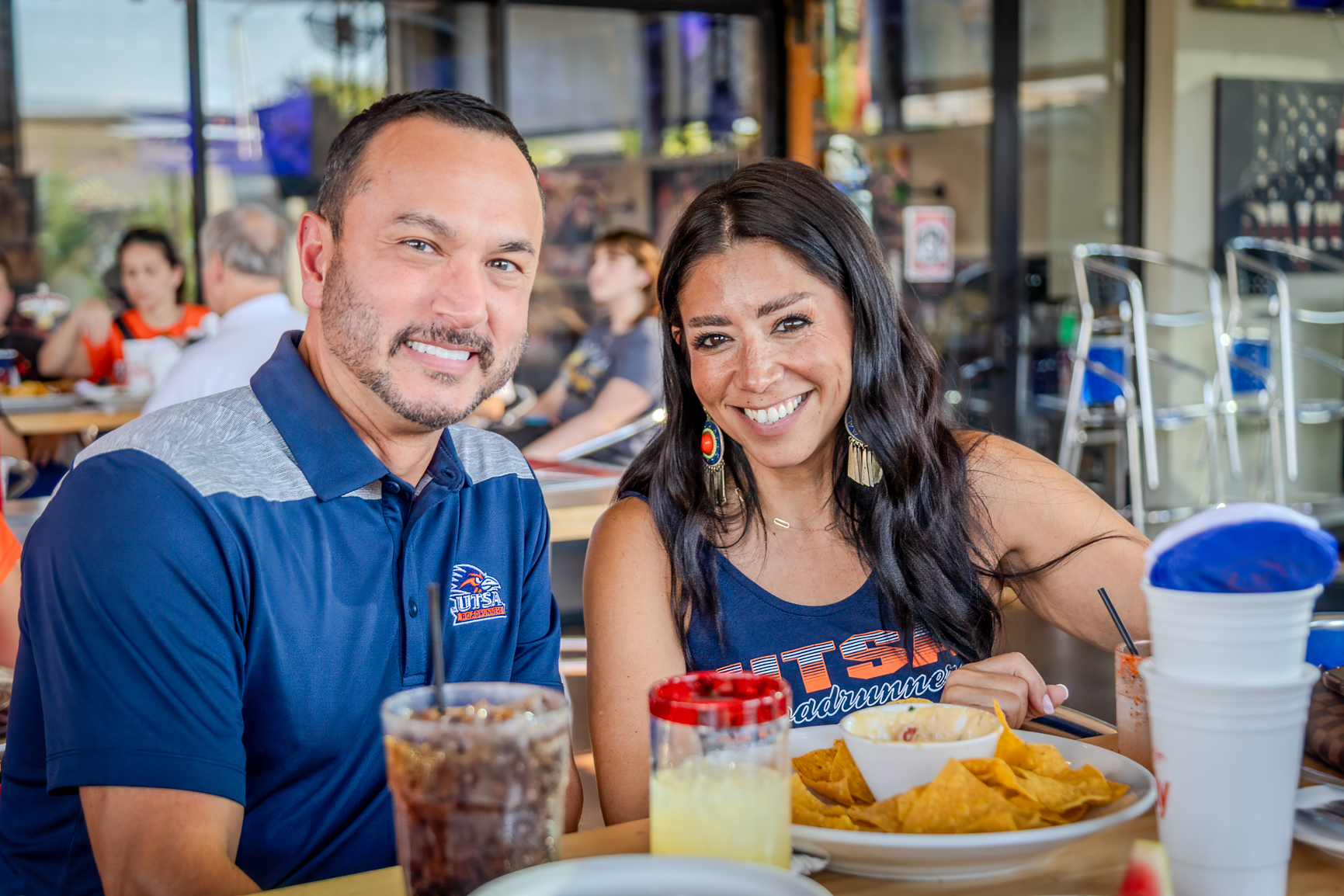 A man and a women smiling at the camera while sitting at a table with food on it, while wearing UTSA shirts