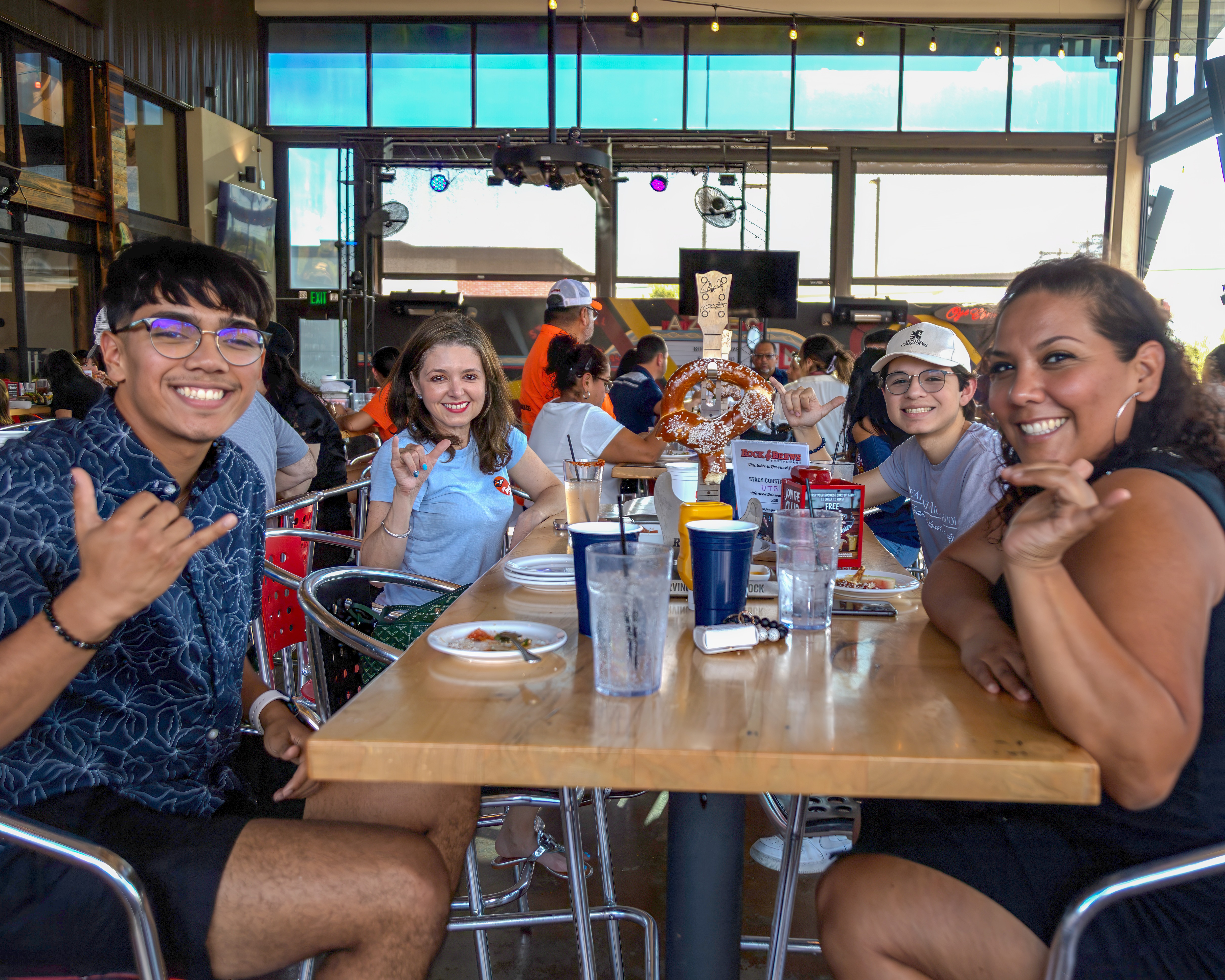 Four people sit around a rectangular table in a crowded restaurant while smiling and holding up the "Birds Up" hand gesture