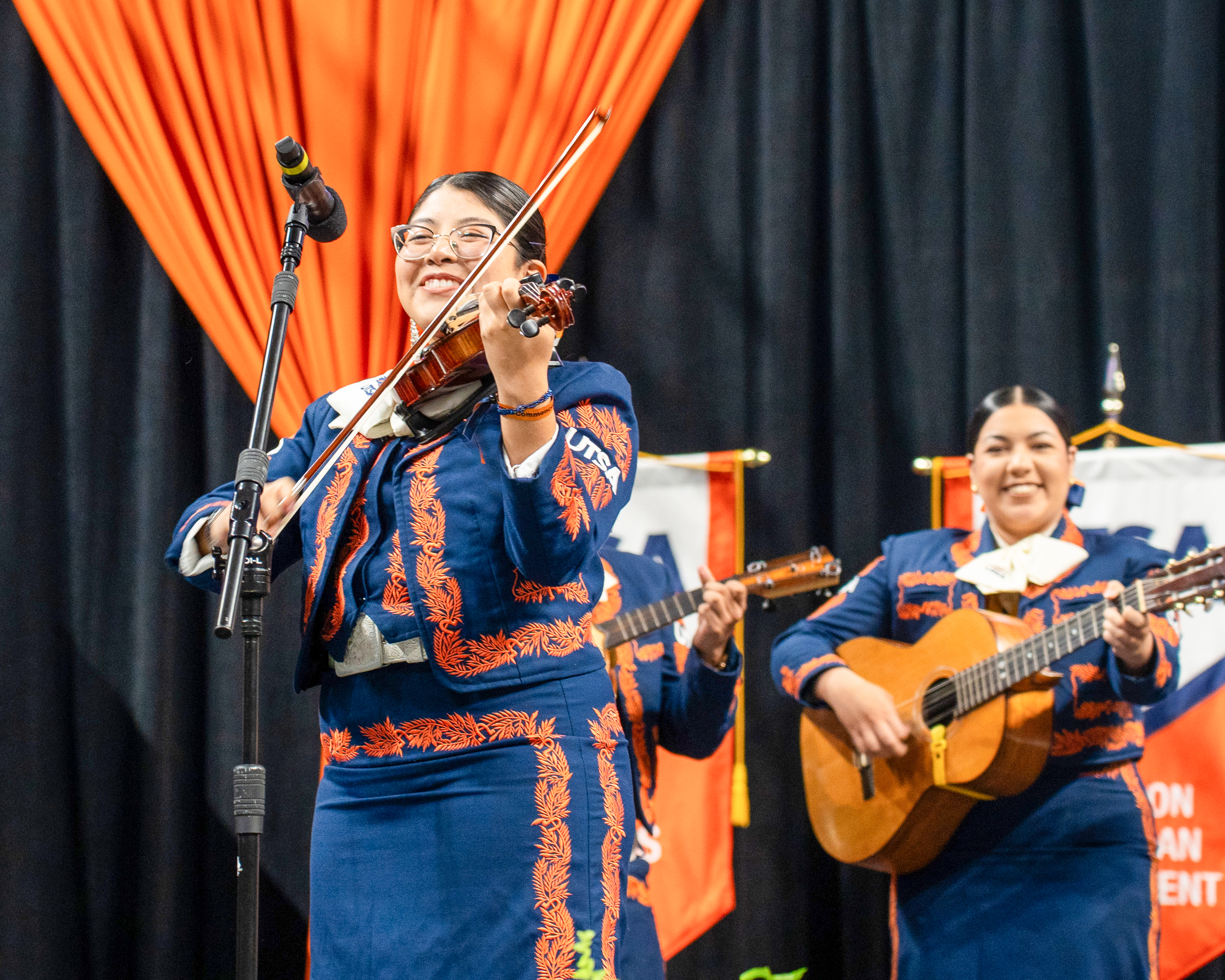 A student dressed in orange and blue Mariachi charro plays the violin while two others play the guitars in the background