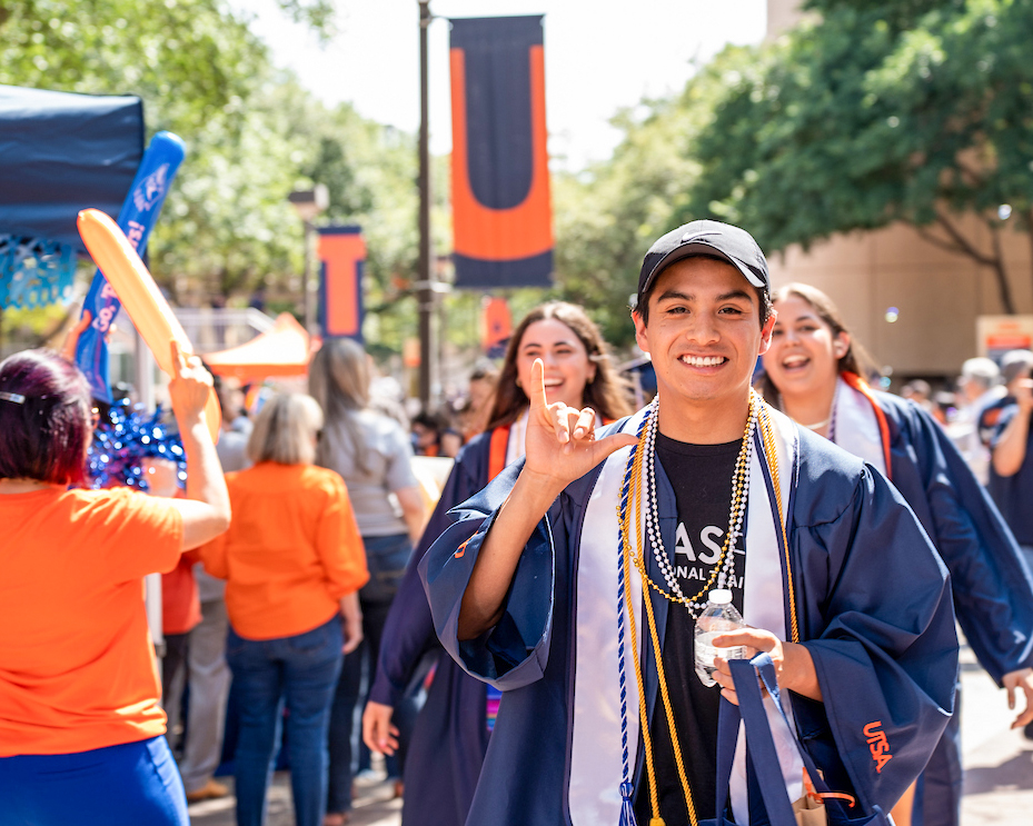 A student walks through a crowd wearing his graduation gown and cords while holding up a "Birds Up" hand gesture.