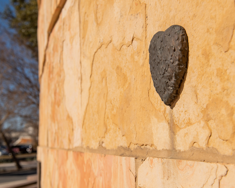 A bronze sculpture of a heart hangs on a stone wall.