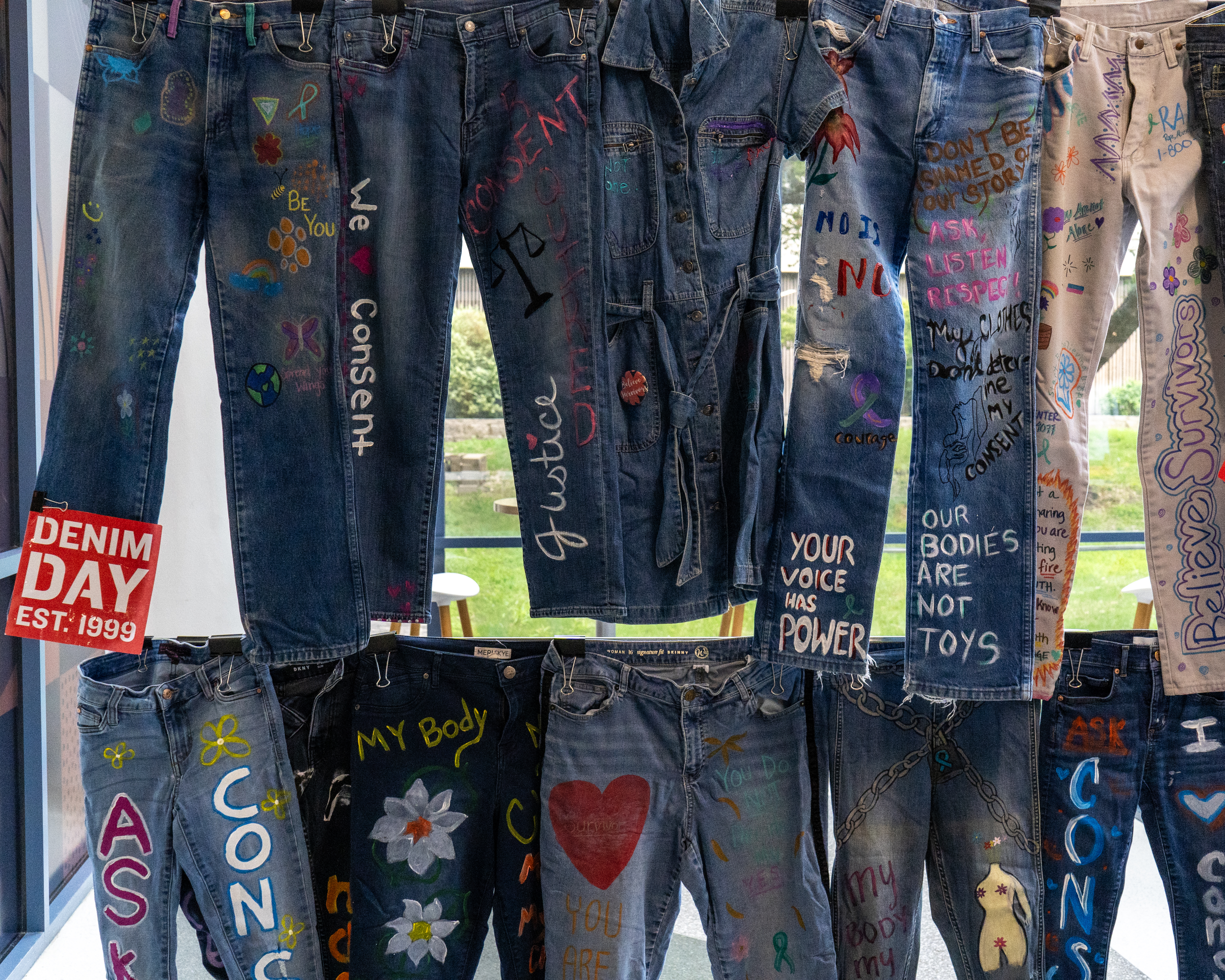 Multiple pairs of jeans hanging in front of a large window. The jeans have writing of words of empowerment for denim day