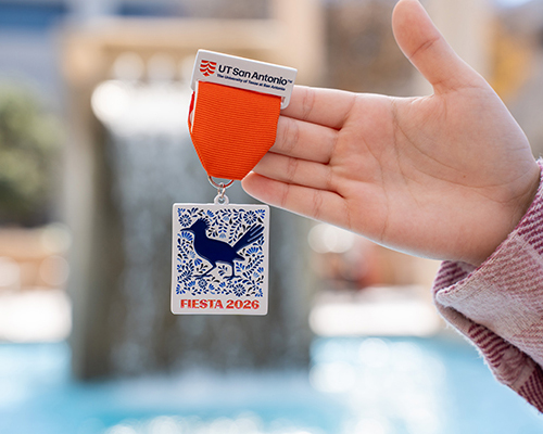 A hand holding a medal that says UT San Antonio across the top with a orange ribbon with a square, white ceramic pendant with blue flower pattern and a blue Roadrunner silhouette with Fiesta 2026 across the bottom.