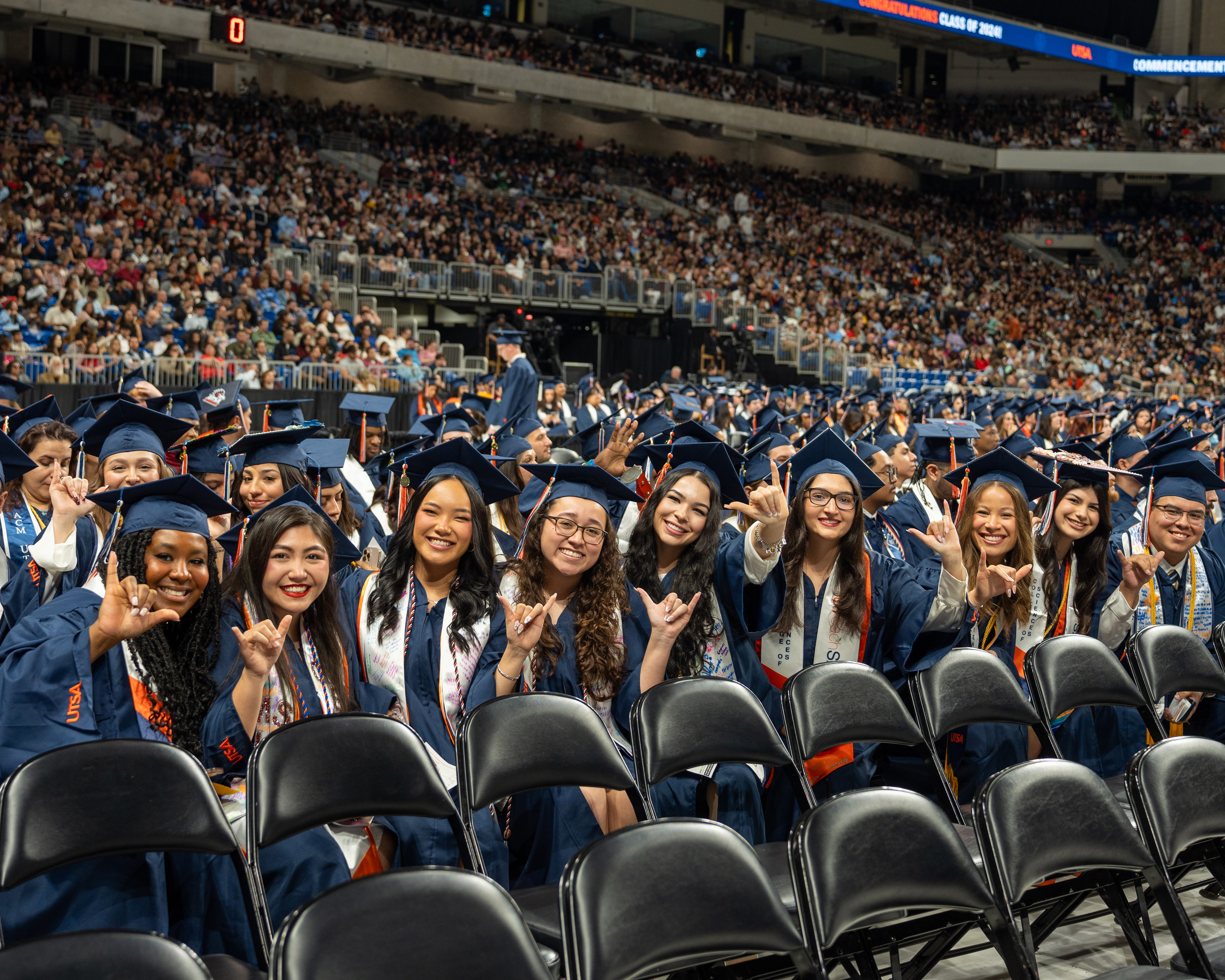 A row of graduates with blue caps and gowns, sit smiling while holding up the "birds up" hand gesture.