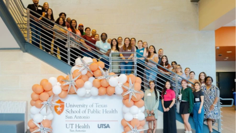 A large group of people standing along a staircase, surrounding a sign that says "University of Texas School of Public Health / UT Health / UT San Antonio and UTSA" with orange and white and star shaped balloons surrounding the sign.