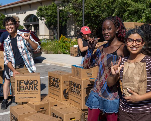 Students holding food band boxes at a food distribution event smile and hold up the "Birds Up" hand gesture.
