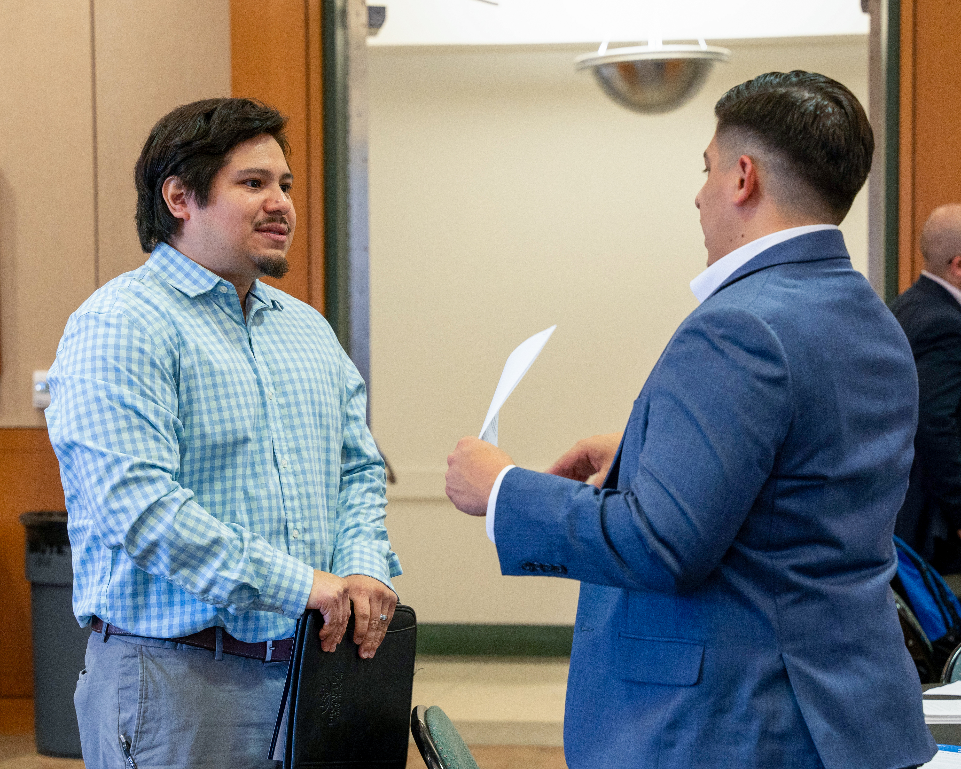 Two people standing in a room talking to each other, one with a piece of paper in his hand and the other with a portfolio in his.