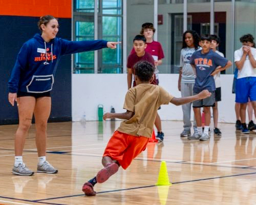 A pre-teen person runs around a yellow cone in a gymnasium with an instructor pointing to the route for the runner to take while a group of participants look on waiting for their turn.