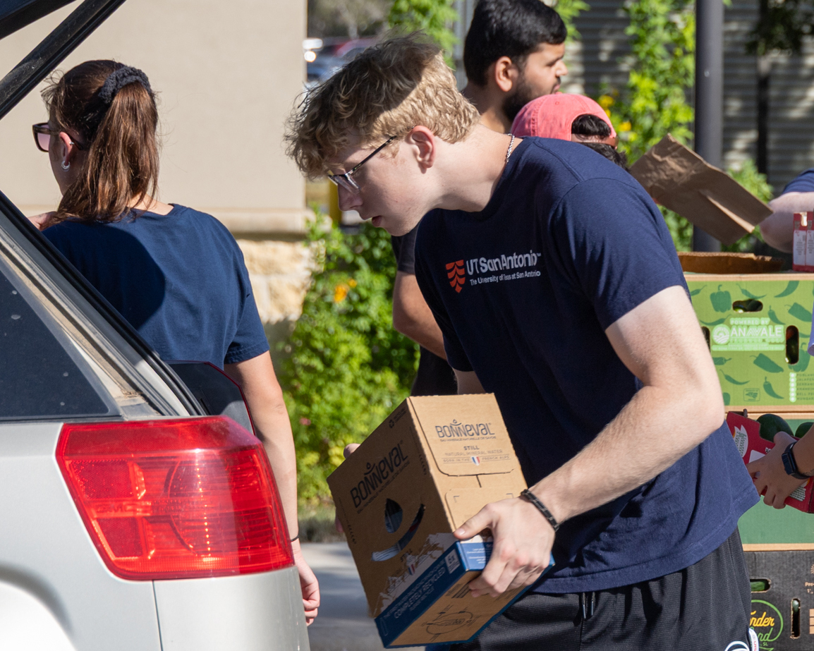A young man loads a box labeled 'Food Bank' into the back of a silver hatchback car.