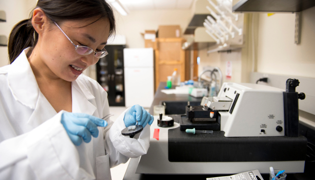 Student with glasses and a pony tail wearing a lab coat and blue gloves analyzes a sample