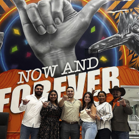 Alumni gathered in front of a mural that says 'Now and Forever' with the roadrunner hand sign