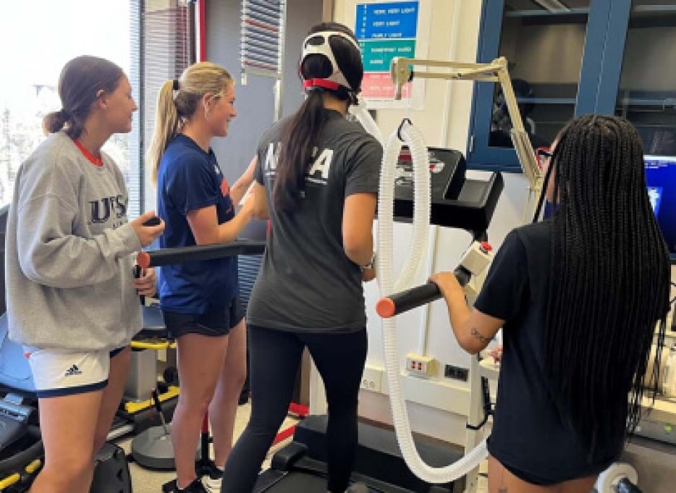 Female on a treadmill wearing an instrument on her head for scientific purposes, while 3 other females look on
