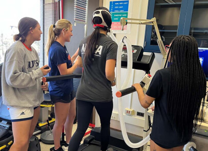 Female on a treadmill wearing an instrument on her head for scientific purposes, while 3 other females look on.