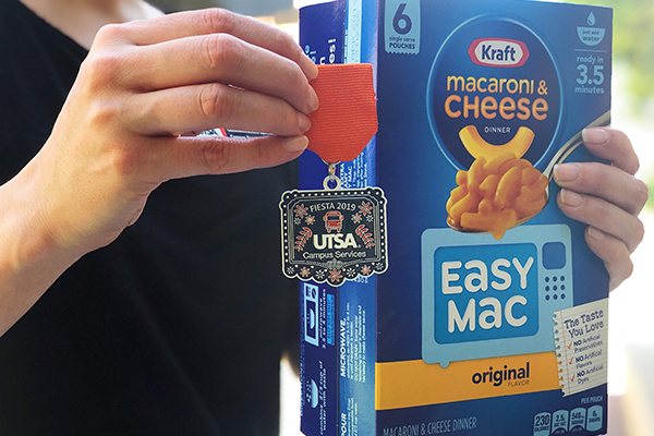 A box of macaroni and cheese is held up next to the Campus Services Fiesta medal.