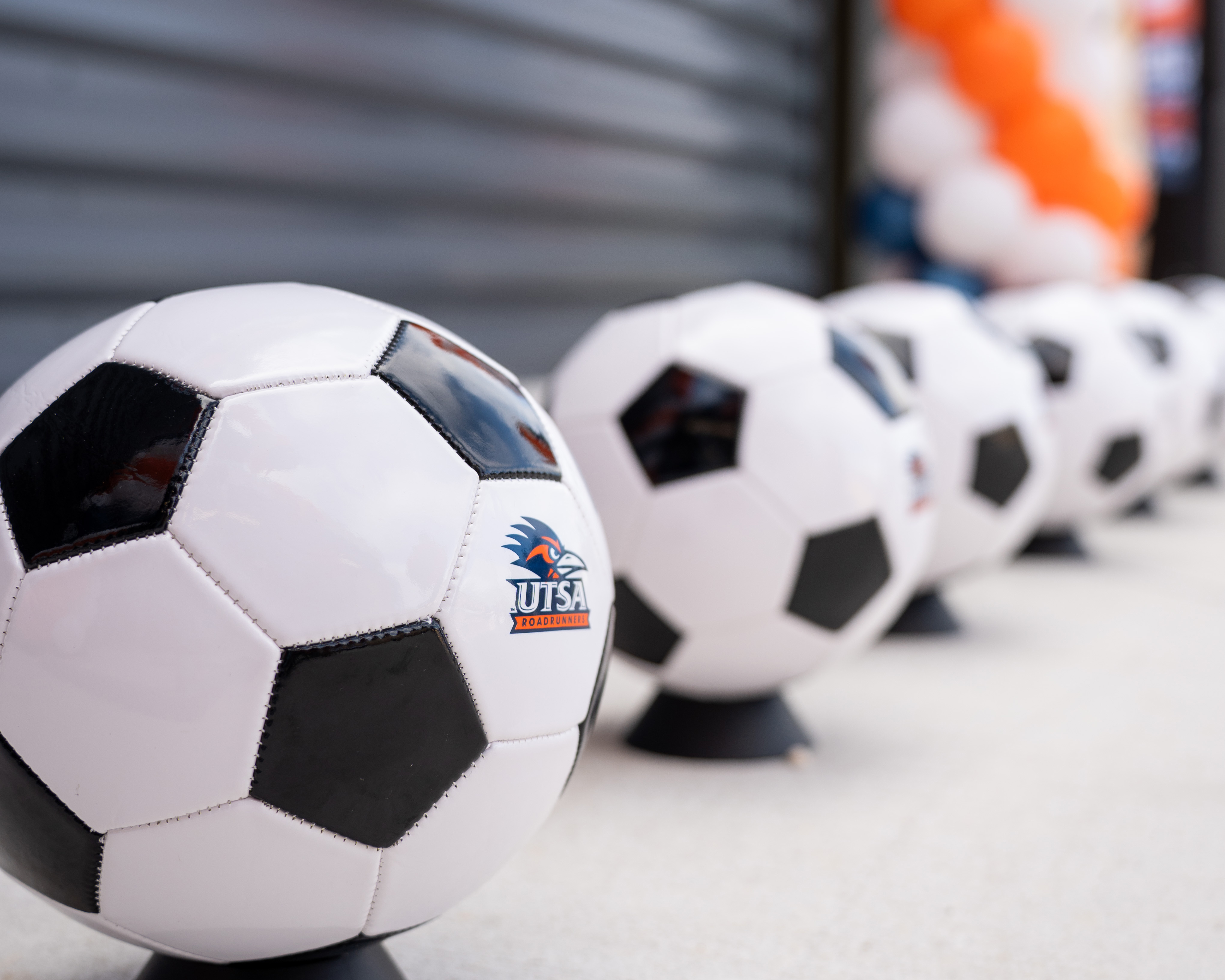 A row of soccer balls blurring into the distance with the a Roadrunner logo on the first ball and colorful balloons in the distance