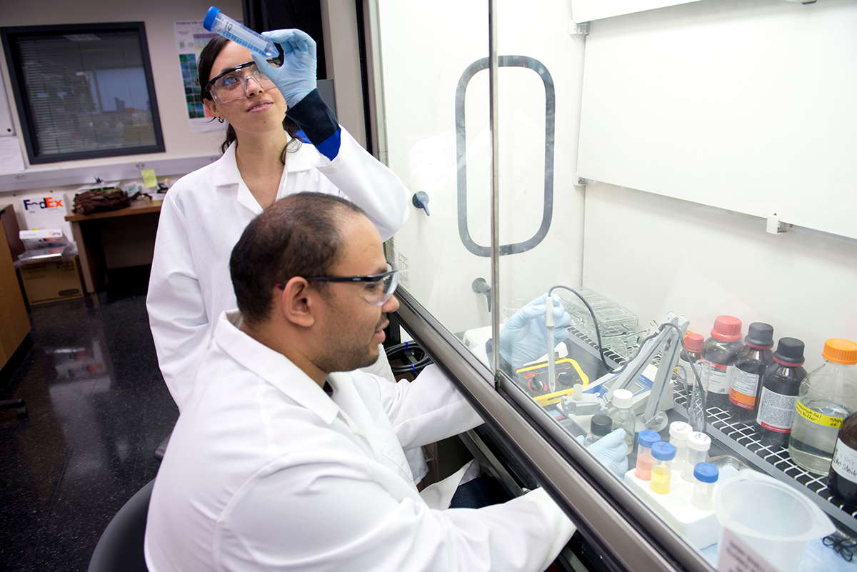 students in lab coats working with chemicals behind glass