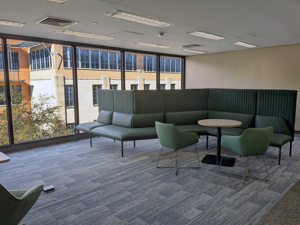 View of JPL collabration space 3rd floor during the day, green furniture for seating and a small work place - two chairs and a small table, a series of windows at the back show Business building during the daytime.