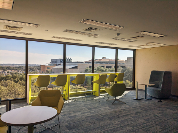 View of JPL collaboration space with seating furniture options including long table and seats in front big windows showing outside of university - trees and building during daytime.