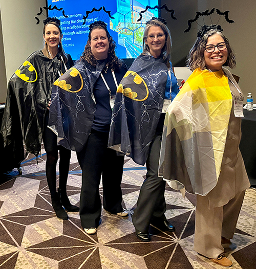 Four Business Affairs leaders standing together, smiling wearing batman costume capes.
