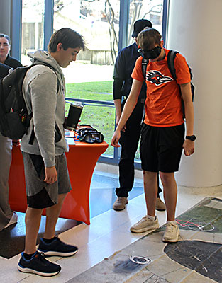 Two male students with backpacks looking down at the floor inside the student union, one in an orange rowdy t-shirt wearing VR goggles. 