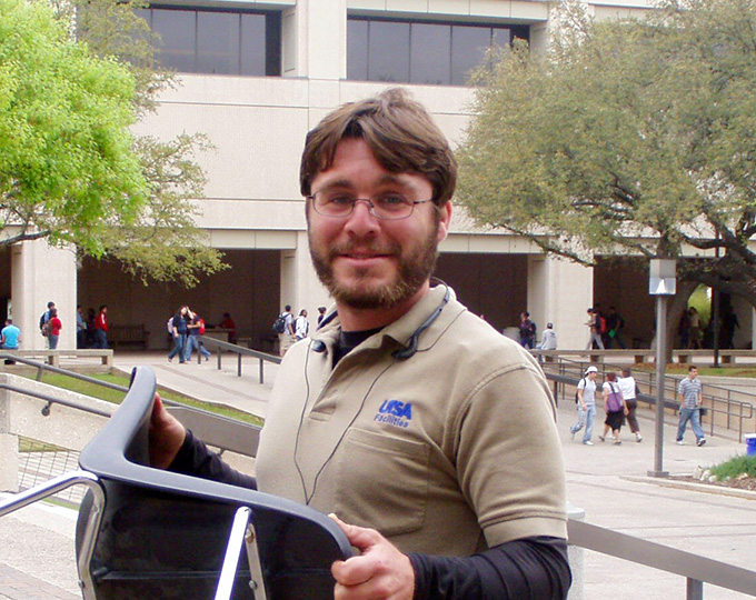 Gregory Peyer smiling and hold a chair outside on campus.