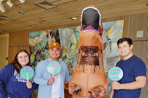 Three people holding I Vote Early signs and a person in a dinosaur costume