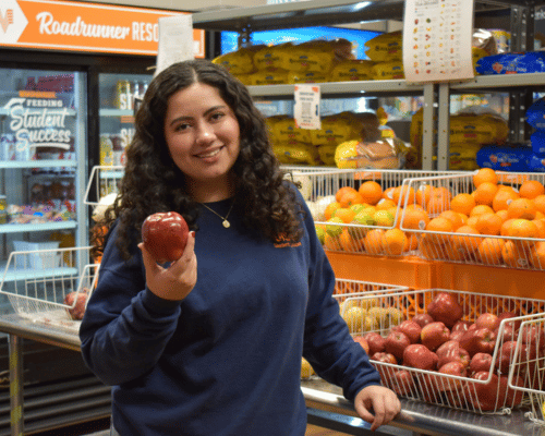 student holding an apple in front of fruit crates in the Roadrunner Pantry