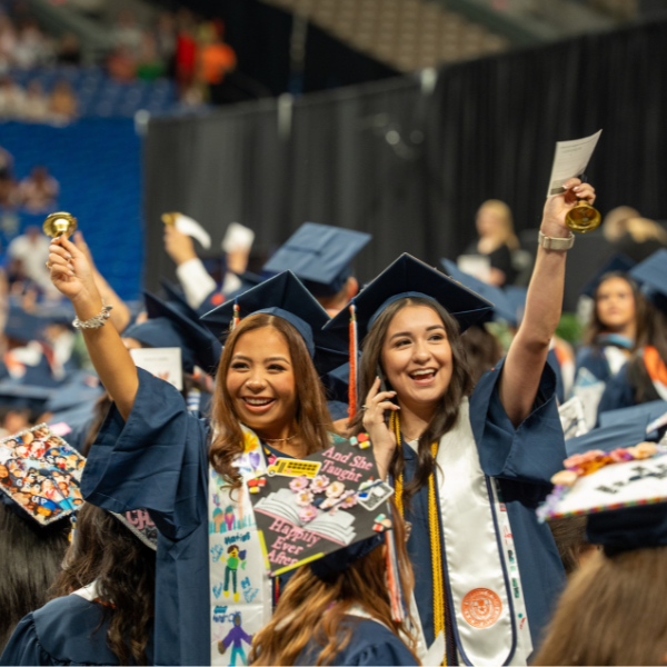 Two students in caps and gowns standing with their arms extended in celebration amid graduating students in the Alamodome