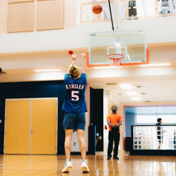 A student in a blue jersey with Kinsler 5 on the back taking a free throw shot while the official watches.