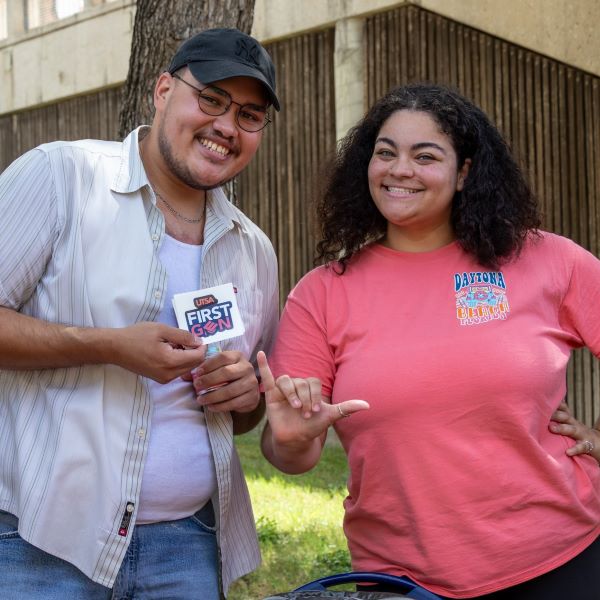 Two students standing behind a table. The person on the left is holding up a First Gen sticker, the person on the right is doing the birds up handsign.