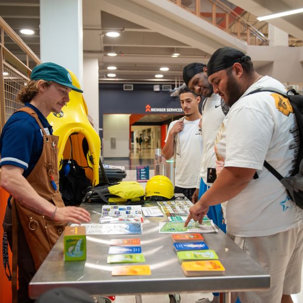 A student stands behind a table with various sheets of paper on it while three studens on the other side of the table read and answer the questions.