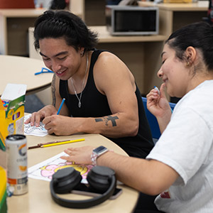 Two people sitting at a table working on coloring book pages. They are smiling and engaging in conversation.
