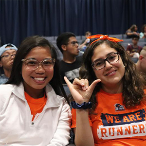 Two students sitting in a theater and smiling. There are more students seated behind them.