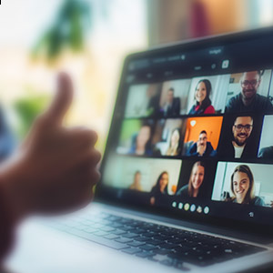 A laptop sitting on a table with a Zoom meeting on the screen. The hand of the laptop owner is giving a thumbs up gesture to the people in the meeting.
